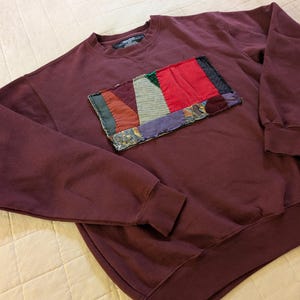 May include: A maroon sweatshirt with a rectangular patchwork design on the chest. The patchwork features various colors including red, green, and purple. The sweatshirt has long sleeves and a crew neck.