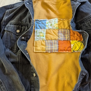 May include: A dark denim jacket layered over a mustard-colored sweatshirt. The sweatshirt features a patchwork design of various fabric patterns and colors, including blue floral, plaid, and orange. The jacket has silver buttons and two chest pockets.