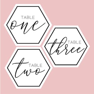 Puede incluir: Tres tarjetas de número de mesa hexagonales blancas con bordes negros y caligrafía negra. Los números son "uno", "dos" y "tres". Cada tarjeta también tiene la palabra "TABLE" impresa encima del número.