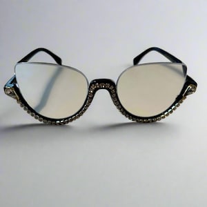 May include: Black cat-eye sunglasses with clear lenses. The frames are embellished with small, sparkling rhinestones along the bottom and at the temples. The top of the frames and the arms are black.