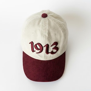 Peut inclure: Casquette de baseball en velours côtelé crème avec une visière bordeaux et le chiffre "1913" brodé sur le devant. La casquette a une couronne arrondie et un bouton sur le dessus. Un accessoire élégant pour un look décontracté.