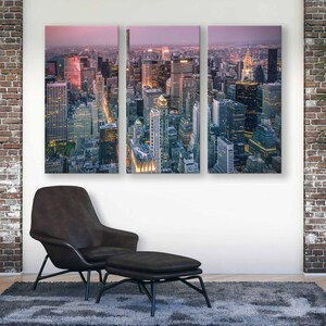Op de afbeelding: Een drieluik canvas print met een hoge hoekweergave van een stadsgezicht in de schemering. De gebouwen zijn verlicht en de lucht heeft een roze en paarse tint. Een moderne zwarte stoel en voetenbank staan voor het kunstwerk.
