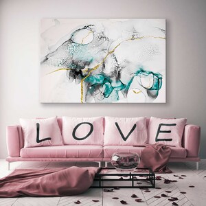 May include: A pink sofa with four cushions spelling out "LOVE" in black lettering. The sofa is in front of a white wall with a large abstract painting featuring teal, black, and gold accents.