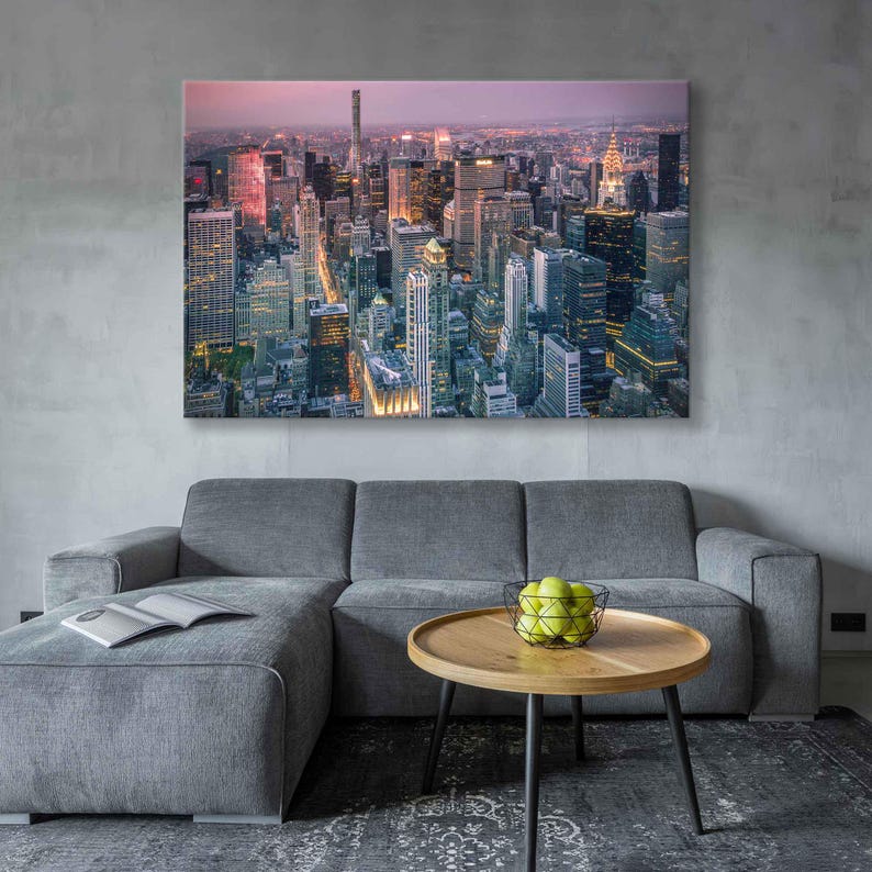 Op de afbeelding: Een groot canvasdoek met een stadsgezicht, met talrijke verlichte wolkenkrabbers. De print hangt boven een grijze hoekbank, met een houten salontafel en een draadmand met groene appels ervoor.