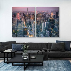 Op de afbeelding: Tweedelige canvas kunst met een hoge hoekweergave van een stadsgezicht in de schemering. Het kunstwerk toont talrijke wolkenkrabbers met verlichte ramen tegen een schemerige roze en paarse lucht. Het kunstwerk wordt weergegeven boven een zwarte bank, salontafel en blauw tapijt.