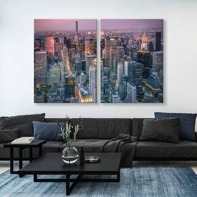 Op de afbeelding: Tweedelige canvas kunst met een hoge hoekweergave van een stadsgezicht in de schemering. Het kunstwerk toont talrijke wolkenkrabbers met verlichte ramen tegen een schemerige roze en paarse lucht. Het kunstwerk wordt weergegeven boven een zwarte bank, salontafel en blauw tapijt.