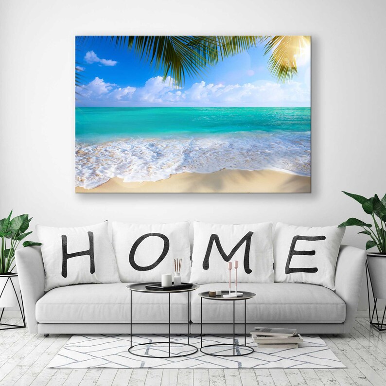 May include: A white sofa with three cushions spelling out "HOME" in black lettering. The sofa is positioned in front of a large canvas print depicting a tropical beach scene with blue water, white sand, and palm trees.