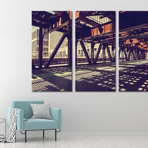 May include: A three-panel canvas print of a city street scene. The image shows a view of a bridge from underneath, with the steel beams and supports in focus. The city skyline is visible in the background, and the sun casts shadows on the ground.