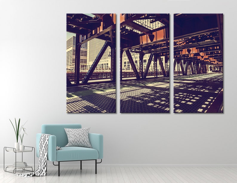 May include: A three-panel canvas print of a city street scene. The image shows a view of a bridge from underneath, with the steel beams and supports in focus. The city skyline is visible in the background, and the sun casts shadows on the ground.