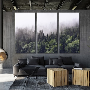 Puede incluir: Una impresi&oacute;n en lienzo de tres paneles que representa una escena de bosque brumoso. La impresi&oacute;n presenta un bosque verde exuberante con &aacute;rboles altos y una niebla suave y et&eacute;rea. La impresi&oacute;n est&aacute; colgada en una pared gris sobre un sof&aacute; gris y dos mesas de caf&eacute; de madera.