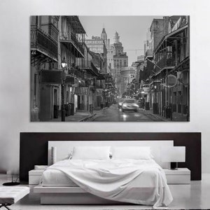 May include: Black and white photograph of a city street scene. Buildings with balconies and awnings line the street. A car is blurred as it moves down the road. The image is displayed above a bed with white linens.
