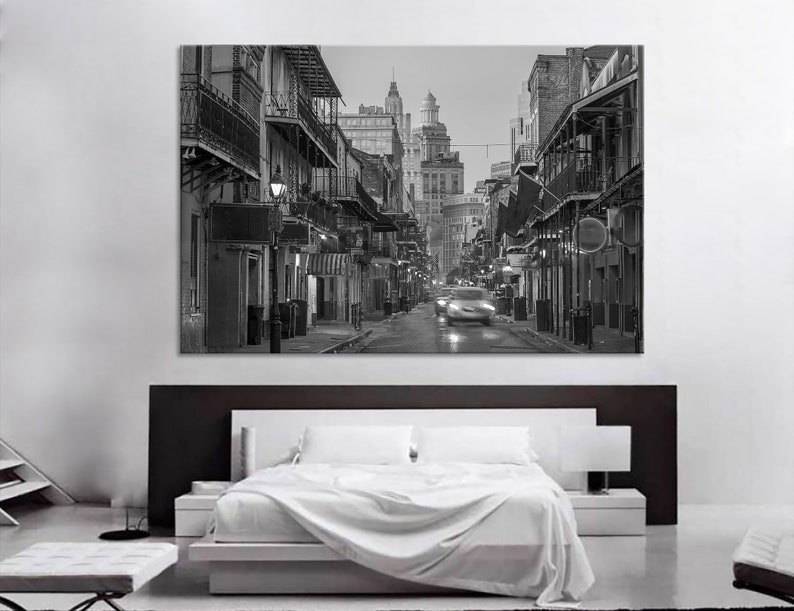 May include: Black and white photograph of a city street scene. Buildings with balconies and awnings line the street. A car is blurred as it moves down the road. The image is displayed above a bed with white linens.