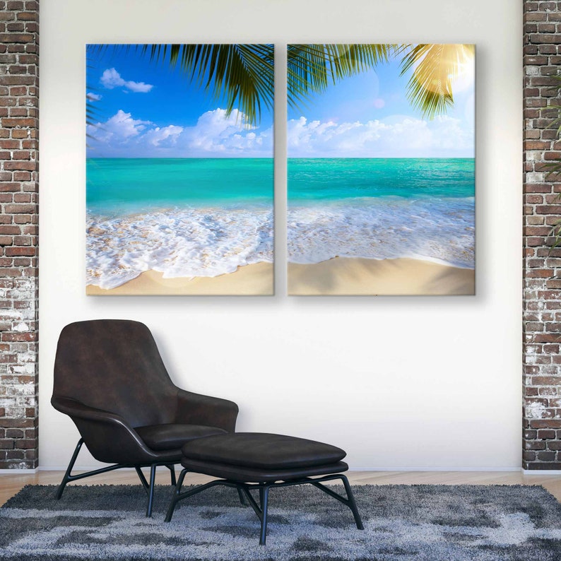 May include: Two canvas prints depicting a tropical beach scene with turquoise water, white sand, and palm trees. The prints are hung on a white wall above a brown leather armchair and footstool.