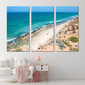 May include: A three-panel canvas print depicting a coastal scene. The artwork showcases a turquoise ocean meeting a sandy beach, with a clear blue sky overhead. The beach is bordered by rocky cliffs and vegetation.