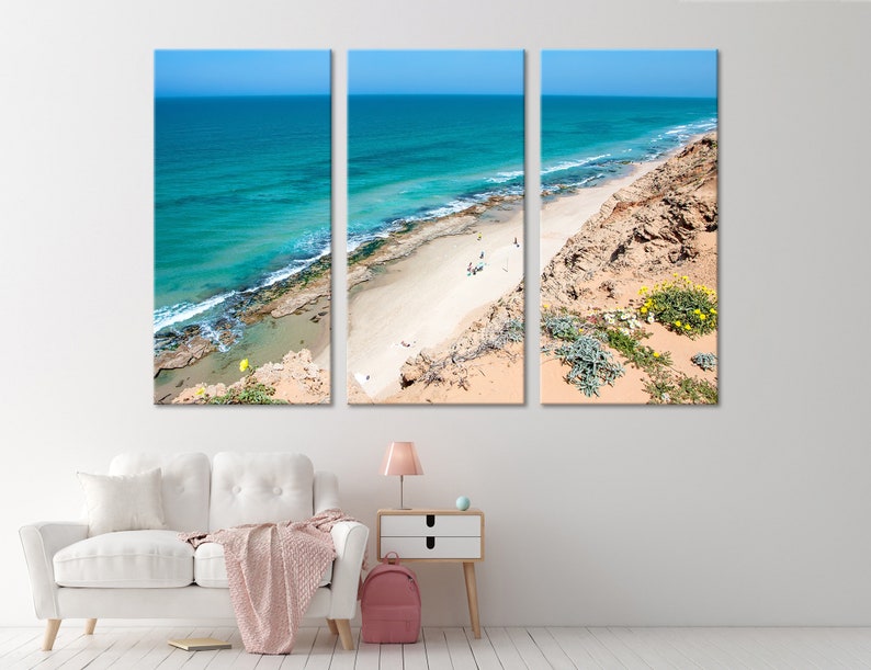May include: A three-panel canvas print depicting a coastal scene. The artwork showcases a turquoise ocean meeting a sandy beach, with a clear blue sky overhead. The beach is bordered by rocky cliffs and vegetation.