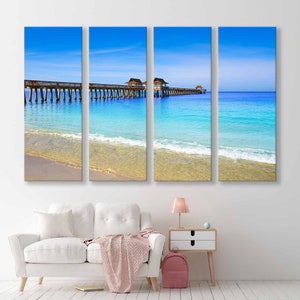 May include: A panoramic view of a wooden pier extending into a turquoise ocean with white sand beach and blue sky. The pier has several small wooden structures on top.
