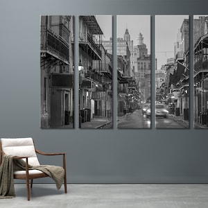 May include: Black and white cityscape art print. The image is a split panel of a street scene with buildings, balconies, and streetlights. Cars are blurred as they move down the wet street. A wooden chair with a blanket is in the foreground.