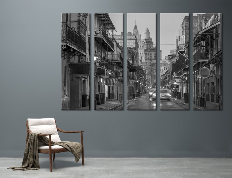 May include: Black and white cityscape art print. The image is a split panel of a street scene with buildings, balconies, and streetlights. Cars are blurred as they move down the wet street. A wooden chair with a blanket is in the foreground.