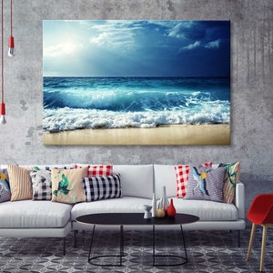 K&ouml;nnte beinhalten: Ein wei&szlig;es Sofa mit bunten Kissen in einem Wohnzimmer. Ein gro&szlig;es Leinwandbild eines blauen Ozeans mit wei&szlig;en Wellen, die an einem Sandstrand brechen, h&auml;ngt an der Wand &uuml;ber dem Sofa.
