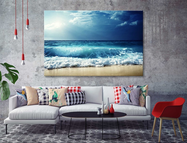 K&ouml;nnte beinhalten: Ein wei&szlig;es Sofa mit bunten Kissen in einem Wohnzimmer. Ein gro&szlig;es Leinwandbild eines blauen Ozeans mit wei&szlig;en Wellen, die an einem Sandstrand brechen, h&auml;ngt an der Wand &uuml;ber dem Sofa.