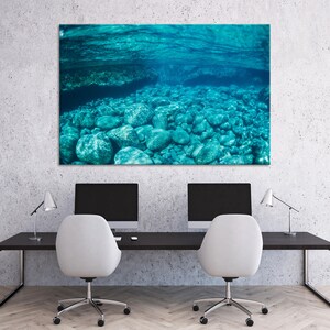 May include: A large rectangular canvas print depicting an underwater scene. The image shows clear turquoise water with rounded rocks on the seabed. The print is mounted on a wall above a black desk with two computer monitors and white chairs.
