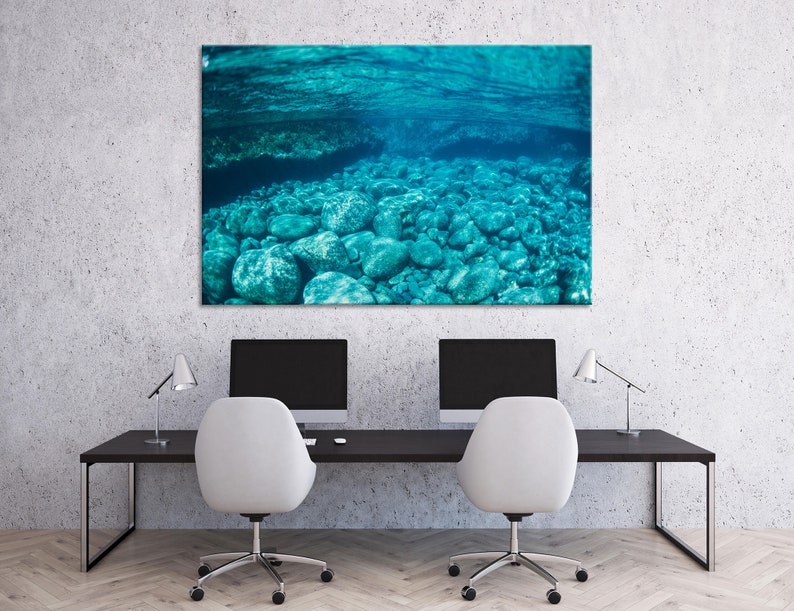 May include: A large rectangular canvas print depicting an underwater scene. The image shows clear turquoise water with rounded rocks on the seabed. The print is mounted on a wall above a black desk with two computer monitors and white chairs.