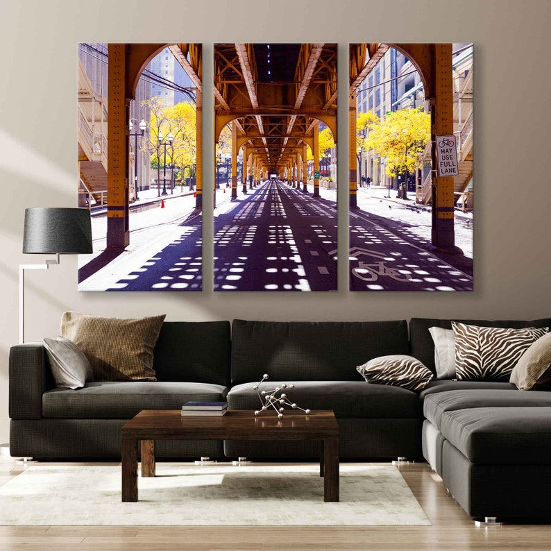 May include: A three-panel canvas print of a city street under a yellow bridge. The street is lined with trees and the sun is shining through the trees, casting shadows on the street. The street is empty except for a few cars and a bicycle lane.
