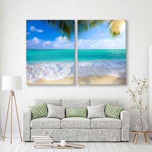 May include: A split canvas print featuring a tropical beach scene with turquoise water, white sand, and palm tree leaves. The image is divided into two panels, each showing a different perspective of the beach.