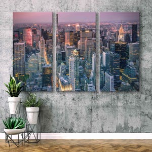 Op de afbeelding: Een drieluik canvas print van een stadsgezicht in de schemering. De afbeelding toont een hoge hoekweergave van talrijke wolkenkrabbers met verlichte ramen en lichten. De lucht gaat over van een zachtroze naar een donkerdere tint, wat een dramatisch contrast cre&euml;ert.