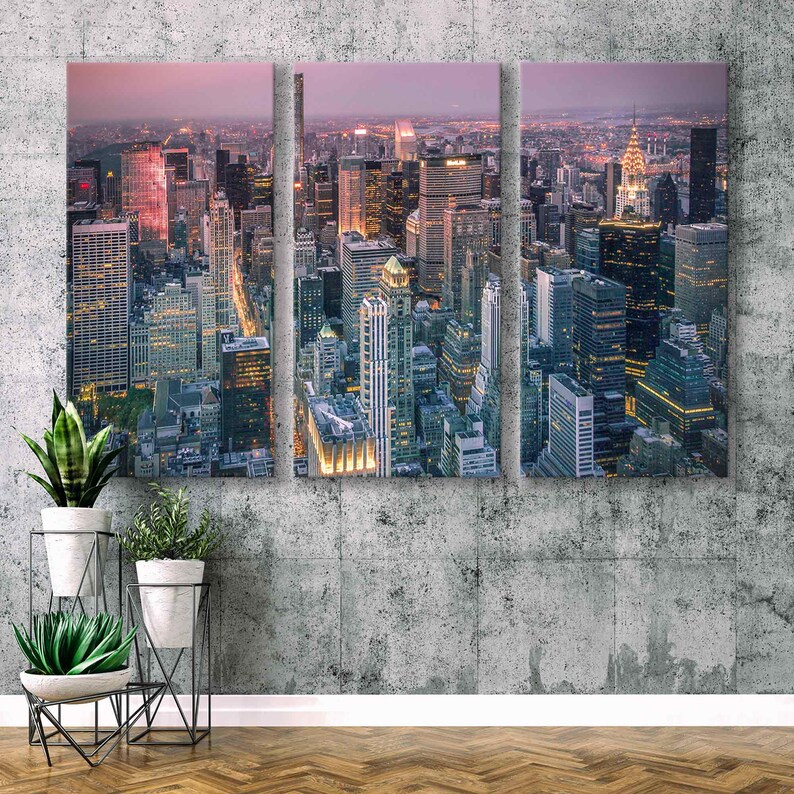 Op de afbeelding: Een drieluik canvas print van een stadsgezicht in de schemering. De afbeelding toont een hoge hoekweergave van talrijke wolkenkrabbers met verlichte ramen en lichten. De lucht gaat over van een zachtroze naar een donkerdere tint, wat een dramatisch contrast cre&euml;ert.