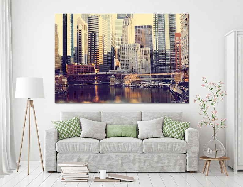 May include: A vintage-style photograph of a cityscape with tall buildings and a river in the foreground. The buildings are mostly brown and gray, with some red accents. The river is calm and reflective, with a bridge crossing it in the distance.