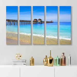 May include: A panoramic view of a wooden pier extending over a turquoise ocean with white sand beach and a blue sky. The pier has several wooden structures on it.