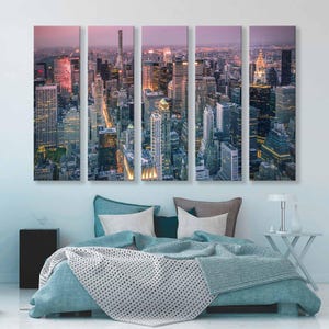 Op de afbeelding: Een canvas print in vijf panelen van een stadsgezicht in de schemering. De afbeelding toont een hoge hoekweergave van talrijke wolkenkrabbers met verlichte ramen, tegen een schemerige roze en paarse lucht. Het kunstwerk wordt weergegeven boven een bed met turquoise beddengoed.