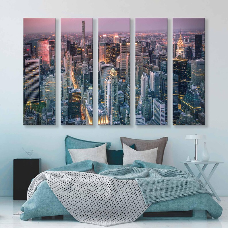 Op de afbeelding: Een canvas print in vijf panelen van een stadsgezicht in de schemering. De afbeelding toont een hoge hoekweergave van talrijke wolkenkrabbers met verlichte ramen, tegen een schemerige roze en paarse lucht. Het kunstwerk wordt weergegeven boven een bed met turquoise beddengoed.