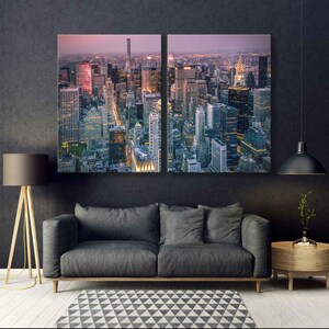Op de afbeelding: Tweedelig canvas kunstwerk met een luchtfoto van een stadsgezicht in de schemering. De gebouwen worden verlicht door warme, gouden lichten, die afsteken tegen de koele, donkere lucht. Het kunstwerk hangt boven een grijze bank, een geometrisch tapijt en een houten vloerlamp.