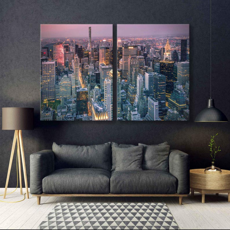 Op de afbeelding: Tweedelig canvas kunstwerk met een luchtfoto van een stadsgezicht in de schemering. De gebouwen worden verlicht door warme, gouden lichten, die afsteken tegen de koele, donkere lucht. Het kunstwerk hangt boven een grijze bank, een geometrisch tapijt en een houten vloerlamp.