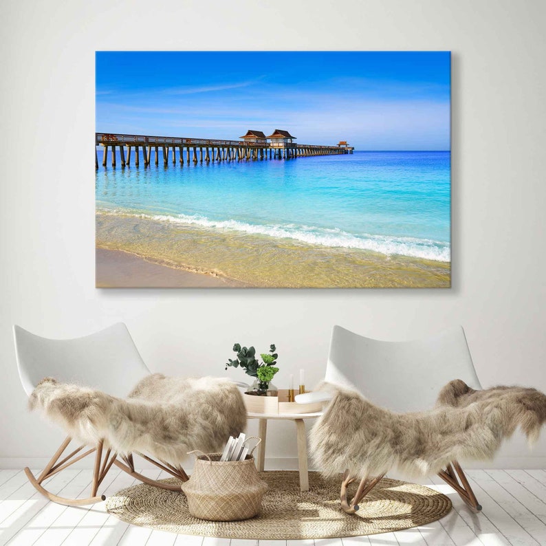 May include: A wooden pier extends over a turquoise ocean with a sandy beach in the foreground. The sky is a bright blue with white clouds.