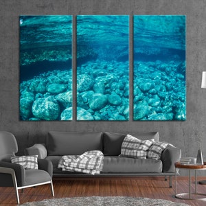 May include: A triptych canvas print depicting an underwater scene. The artwork showcases a view of a riverbed with numerous rounded stones in shades of teal and blue, viewed from below the water's surface. The water above is visible, creating a serene and natural aesthetic.