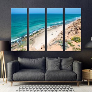 May include: Four-panel canvas art print depicting a coastal scene. The artwork showcases a turquoise ocean, a sandy beach, and a rocky cliff under a clear blue sky. The panels are displayed above a gray sofa in a modern living room setting.