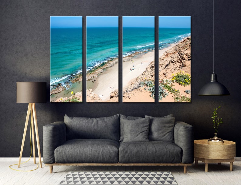 May include: Four-panel canvas art print depicting a coastal scene. The artwork showcases a turquoise ocean, a sandy beach, and a rocky cliff under a clear blue sky. The panels are displayed above a gray sofa in a modern living room setting.