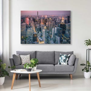 Op de afbeelding: Een groot canvasdoek met een stadsgezicht in de schemering, met talrijke wolkenkrabbers met verlichte ramen. De print hangt boven een grijze bank met decoratieve kussens en een witte salontafel met planten.