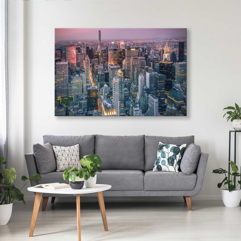 Op de afbeelding: Een groot canvasdoek met een stadsgezicht in de schemering, met talrijke wolkenkrabbers met verlichte ramen. De print hangt boven een grijze bank met decoratieve kussens en een witte salontafel met planten.