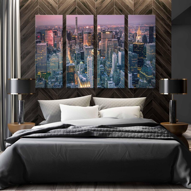 Op de afbeelding: Een vierdelige canvas print van een stadsgezicht in de schemering, met verlichte wolkenkrabbers. Het kunstwerk hangt boven een bed met witte kussens en een grijze dekbedovertrek. Twee zwarte lampen met gouden voetstukken flankeren het bed.