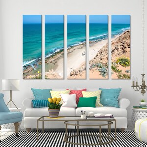 May include: Five-panel canvas art print depicting a coastal scene. The artwork showcases a turquoise ocean meeting a sandy beach under a clear blue sky. The beach is bordered by a rocky cliff with sparse vegetation.