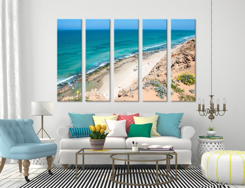 May include: Five-panel canvas art print depicting a coastal scene. The artwork showcases a turquoise ocean meeting a sandy beach under a clear blue sky. The beach is bordered by a rocky cliff with sparse vegetation.