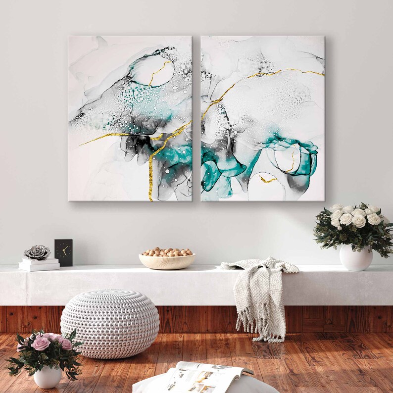 May include: Two abstract paintings with a white background, teal and black swirls, and gold accents. The paintings are hanging on a white wall above a white shelf with a bowl of nuts, a white footstool, and a white throw blanket.