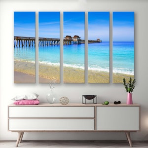 May include: A five-panel canvas print depicting a wooden pier extending into a turquoise ocean with whitecaps. The pier has several small, wooden structures on it. The beach is sandy and the sky is blue.