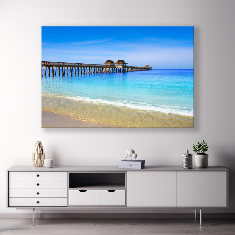 May include: A photograph of a wooden pier extending into a turquoise ocean with a sandy beach in the foreground. The sky is a bright blue with white clouds.
