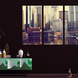 May include: A three-panel canvas print featuring a cityscape with tall buildings, a river, and a bridge. The buildings are a mix of modern and traditional styles, and the river is reflecting the light of the setting sun. The print is in a vintage style, with a warm color palette.