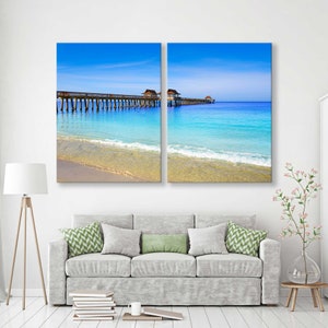 May include: A split canvas print of a wooden pier extending into a turquoise ocean with a sandy beach in the foreground. The sky is a bright blue with white clouds.